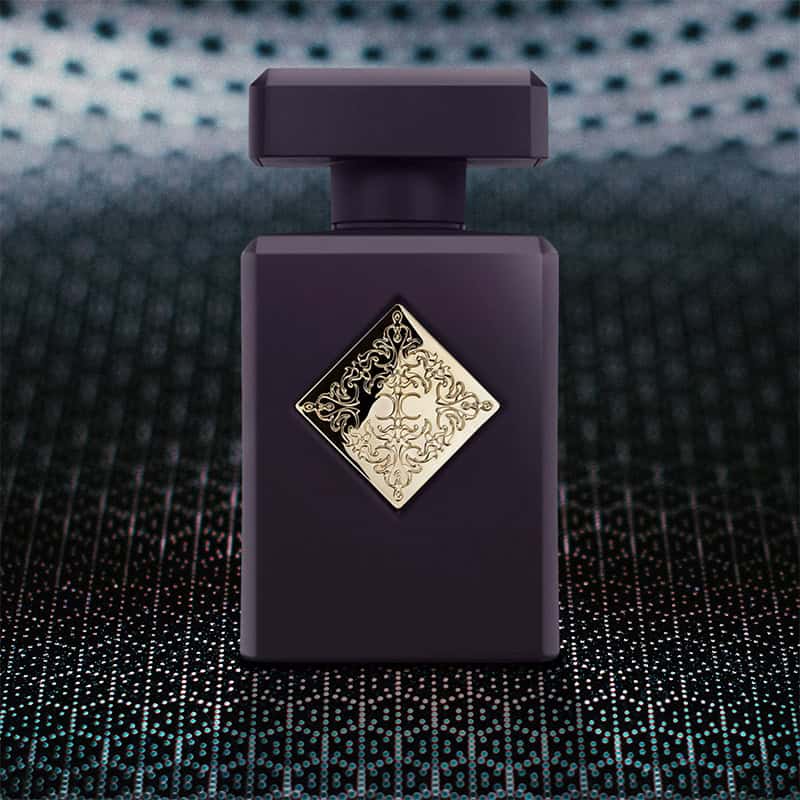 A close-up of the front face of a bottle of Initio Parfums Atomic Rose. The bottle is sitting on a textured grey-colored hexagonal-patterned surface and there is a blurred hexagonal-patterned background.