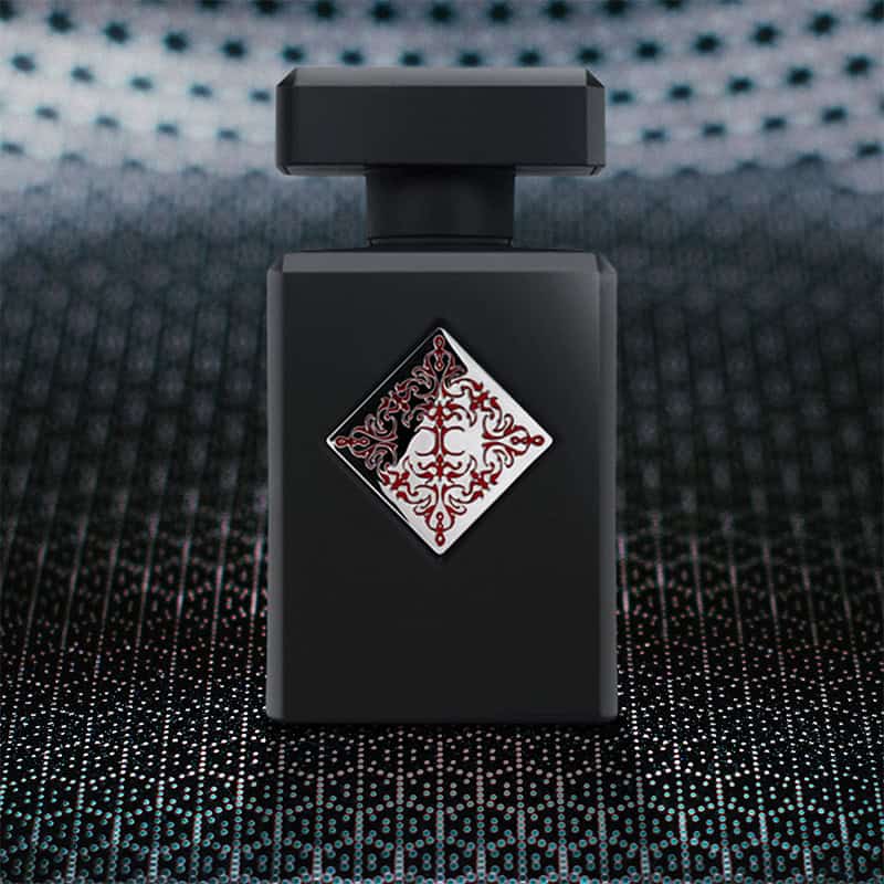 A close-up of the front face of a bottle of Initio Parfums Blessed Baraka. The bottle is sitting on a textured grey-colored hexagonal-patterned surface and there is a blurred hexagonal-patterned background.