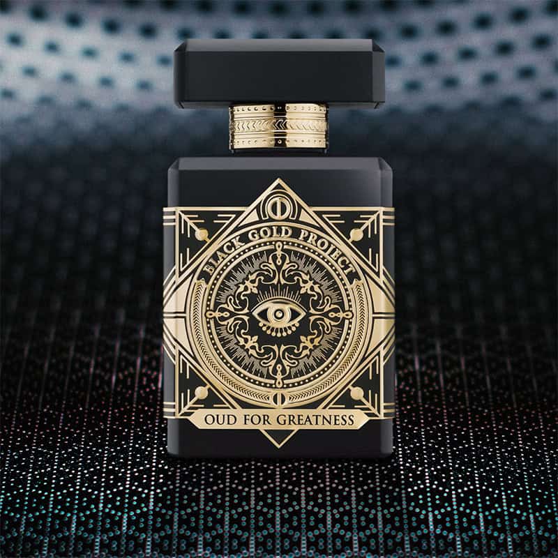 A close-up of the front face of a bottle of Initio Parfums Oud For Greatness. The bottle is sitting on a textured grey-colored hexagonal-patterned surface and there is a blurred hexagonal-patterned background.