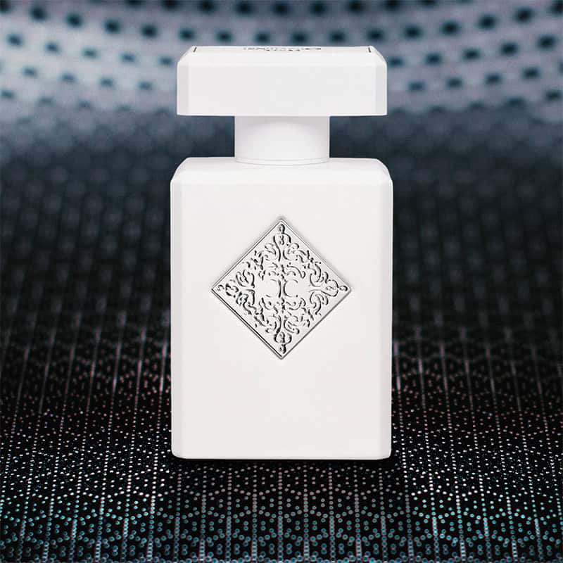 A close-up of the front face of a bottle of Initio Parfums Rehab. The bottle is sitting on a textured grey-colored hexagonal-patterned surface and there is a blurred hexagonal-patterned background.