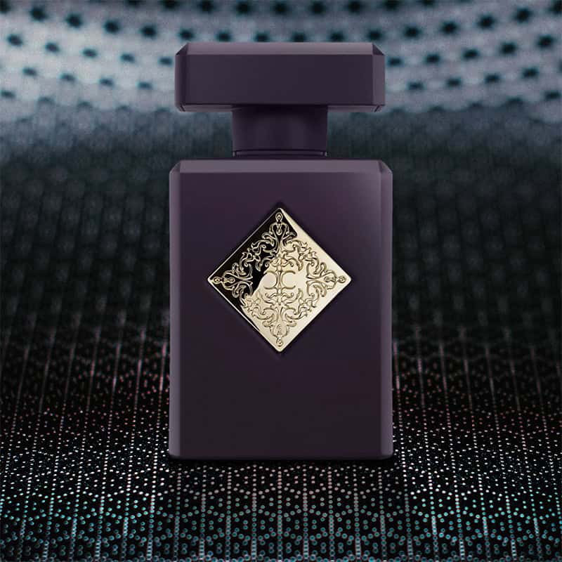 A close-up of the front face of a bottle of Initio Parfums Side Effect. The bottle is sitting on a textured grey-colored hexagonal-patterned surface and there is a blurred hexagonal-patterned background.