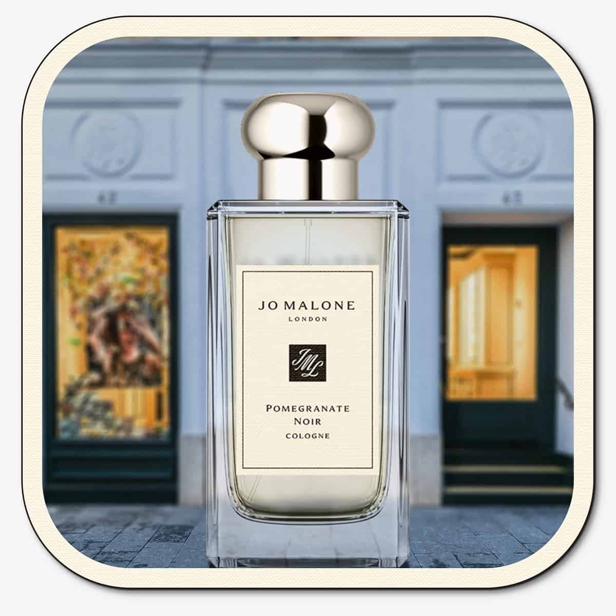 A close-up of the front face of a bottle of Jo Malone Pomegranate Noir Cologne. The bottle is placed on the ground with the Jo Malone global flagship storefront in 
London in the background.