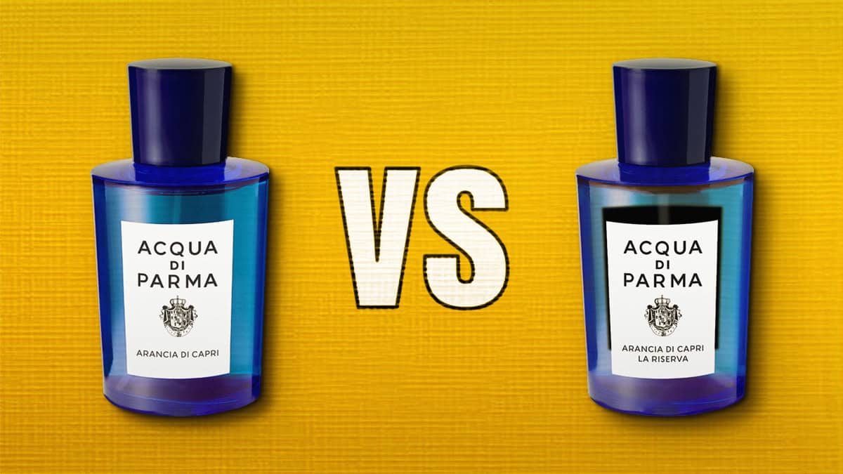 A close-up comparison of the front faces of a bottle of Acqua Di Parma Arancia di Capri and a bottle of Acqua di Parma Arancia di Capri La Riserva, which is placed to the right of it. The 2 bottles are placed on their side next to each other, label side up, and are resting on a yellow-colored textured surface. Between the 2 bottles is a white-colored VS text printed on the background surface.