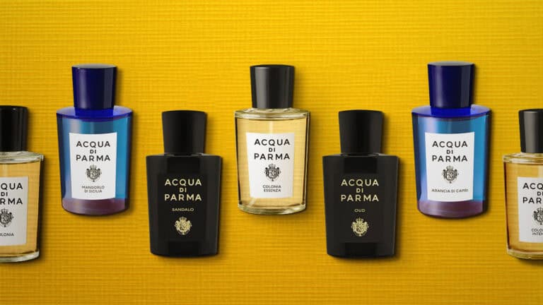 7 Acqua di Parma fragrance bottles that are evenly lined up next to each other in a row in a staggered high-low pattern. All the bottles are placed on their side, label side up, and are laid out on a yellow-colored textured surface. From left to right, the fragrances are Colonia, Mandorlo di Sicilia, Sandalo, Colonia Essenza, Oud, Arancia di Capri, and Colonia Intensa.