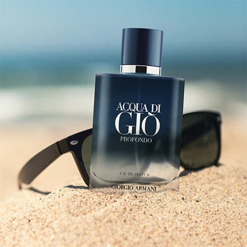 A close-up of an updated new-look Giorgio Armani Acqua Di Gio Profondo Eau De Parfum bottle. The bottle is sitting in the sand on the beach, and is placed in front of a pair of sunglasses with the sea horizon in the background.